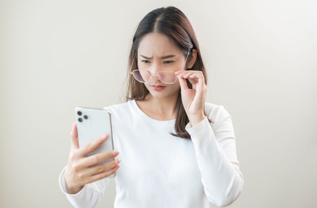 Woman squinting at phone screen and adjusting glasses, experiencing blurry vision from astigmatism.